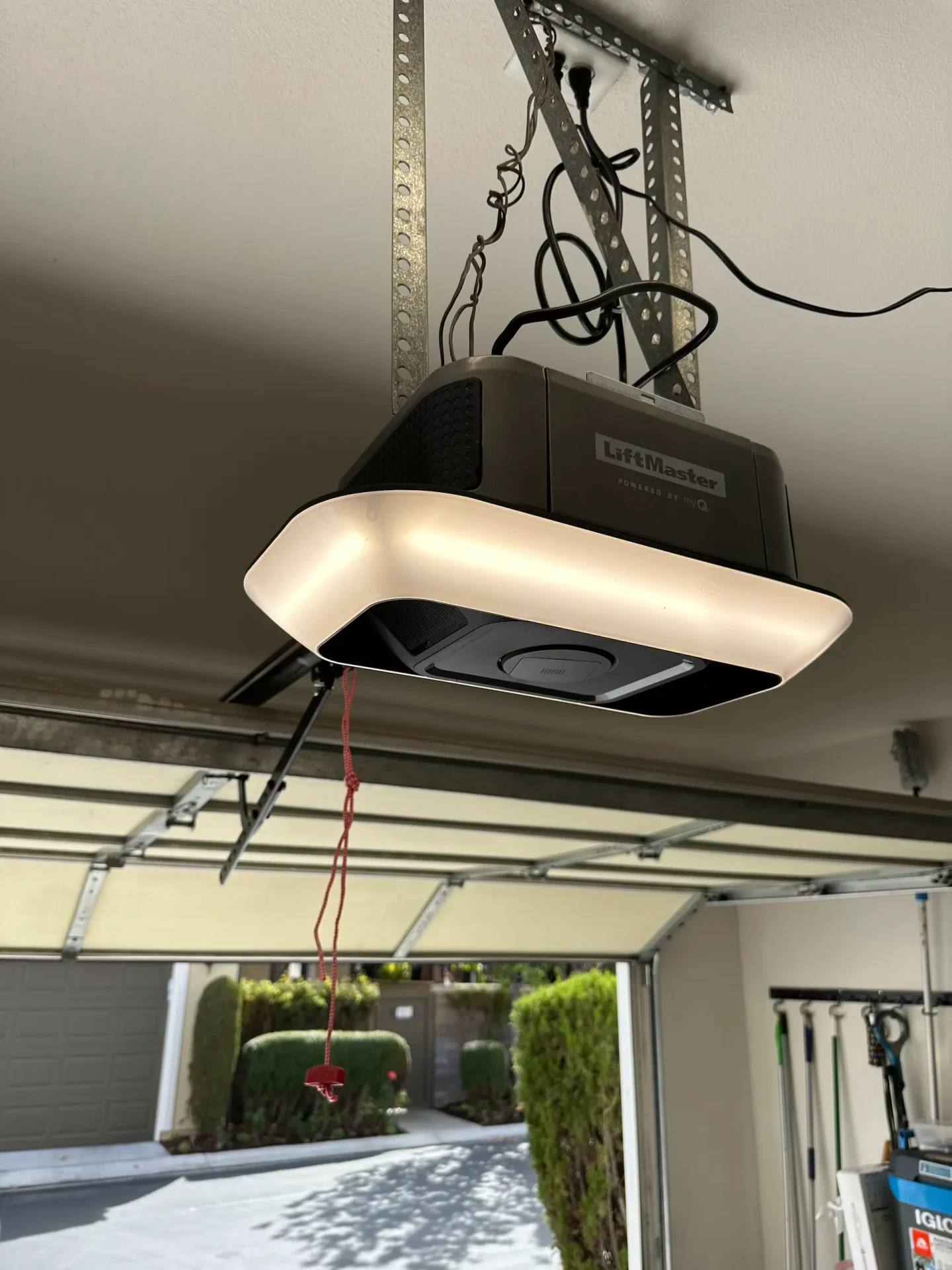 A LiftMaster garage door opener with an illuminated light is mounted on the ceiling, featuring a red emergency release cord hanging down. The open garage door reveals a driveway with greenery—ideal for those seeking reliable garage door service.
