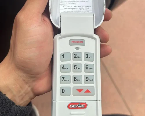 A hand holds a Genie garage door keypad with the cover flipped open, revealing a numeric keypad and “PROGRAM” button at the top. The Genie logo is visible near the bottom—perfect for those seeking reliable garage door repair Orange County services.